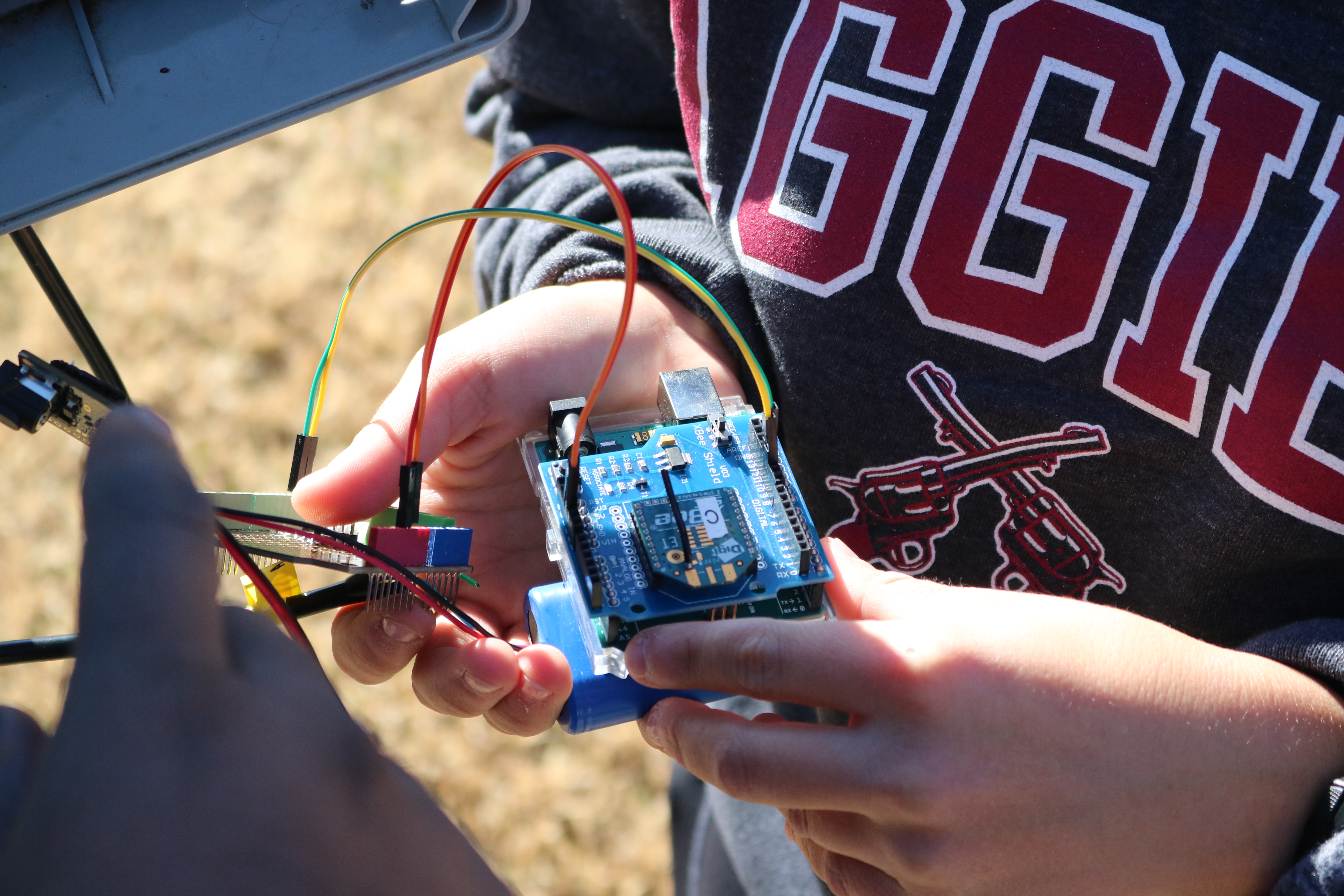 Hands holding a small Arduino-style circuit board with jumper wires connected, outdoors.